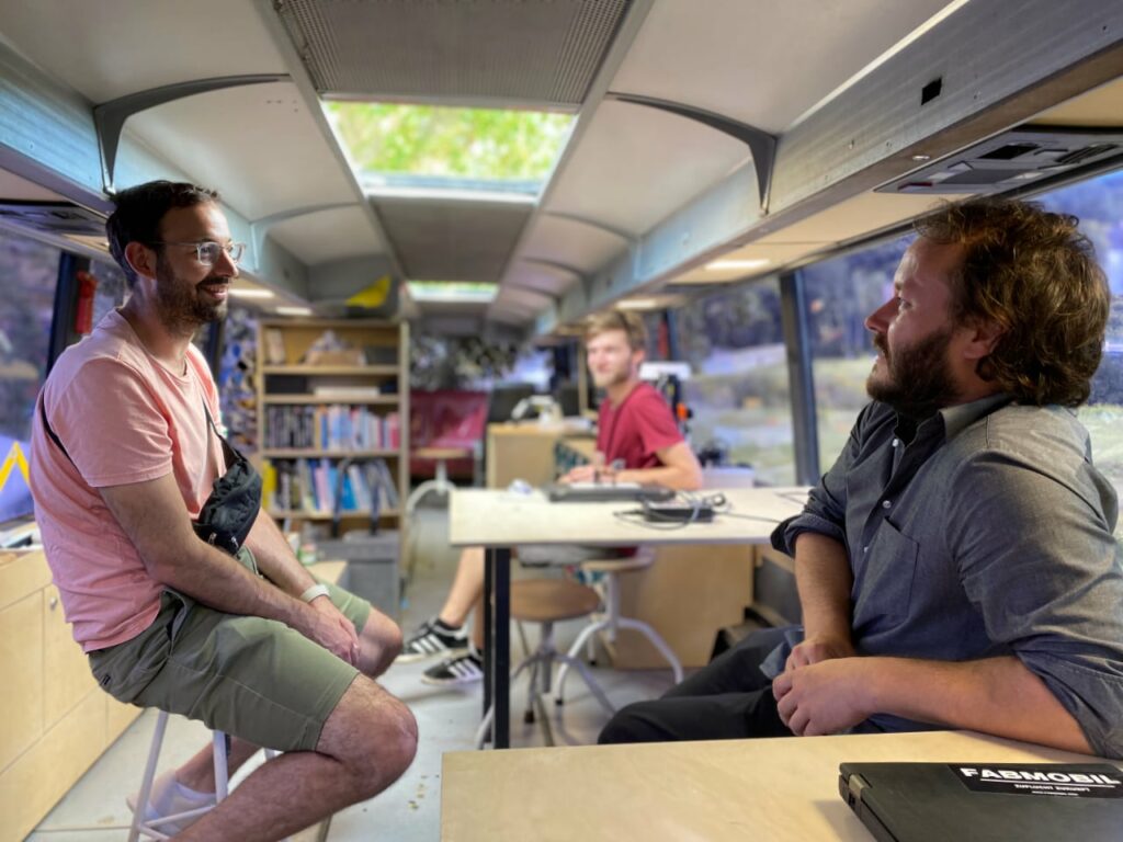 Daniel sitzt gemeinsam mit den Mitarbeiter:innen im Fabmobil und sie unterhalten sich angeregt. Im Hintergrund sieht man die Ausstattung des Buses: ein Bücherregal, einen Arbeitstisch und technische Geräte.