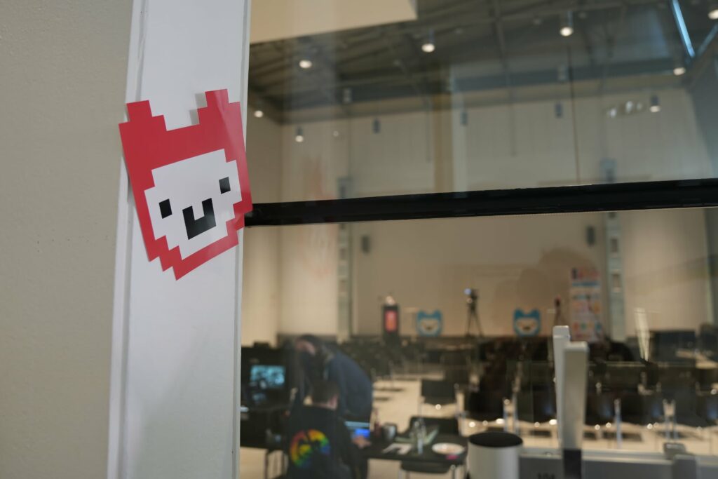Blick durch eine Glasfront in den Vortragssaal mit rotem Jugend hackt Logo, einem Alpaka, links am Fenster