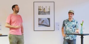 Der Fotograf Wieland Götzler und Daniel Gerber sind auf dem Bild bei der Ausstellungseröffnung zu sehen. Im Zentrum des Bildes sind Fotografien von einem Star und einem Grünspecht zu sehen.