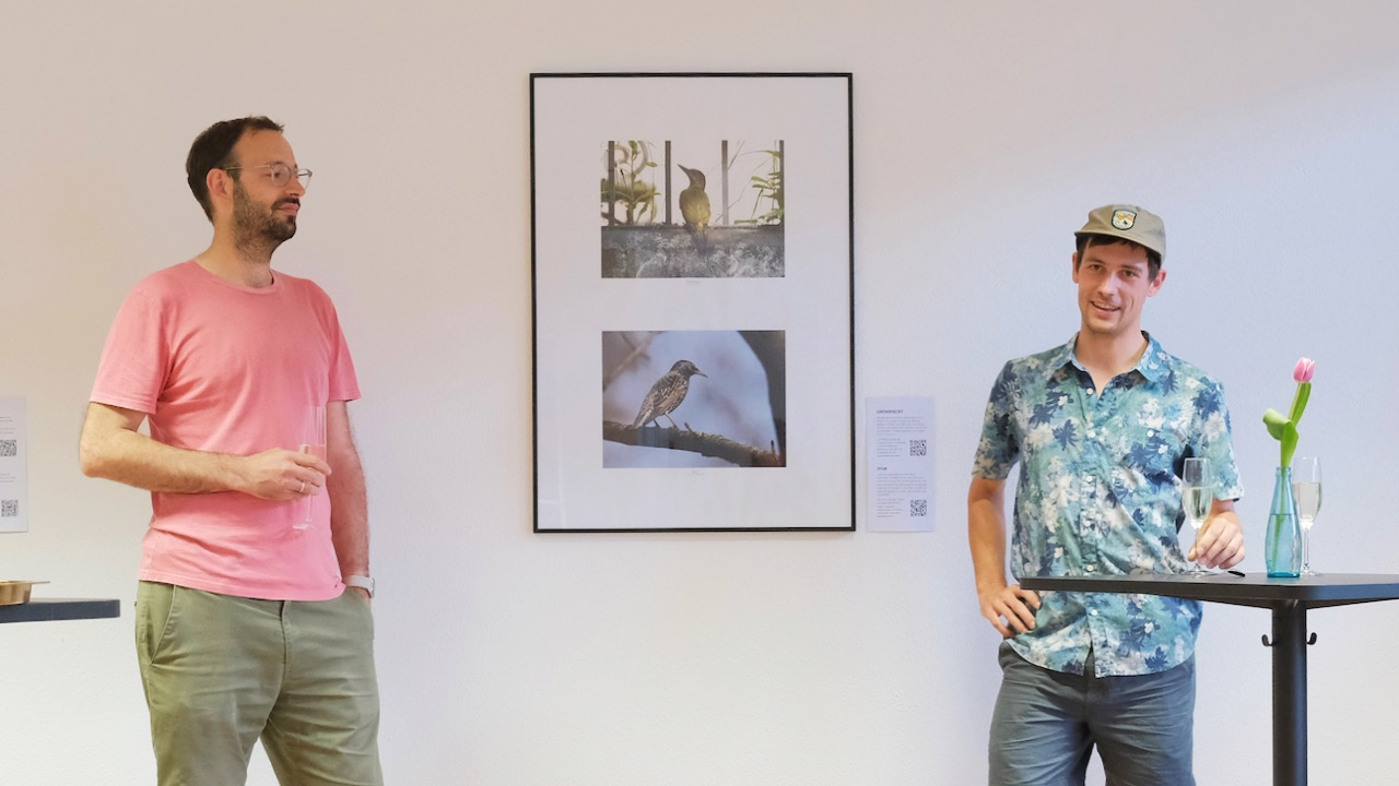Der Fotograf Wieland Götzler und Daniel Gerber sind auf dem Bild bei der Ausstellungseröffnung zu sehen. Im Zentrum des Bildes sind Fotografien von einem Star und einem Grünspecht zu sehen.