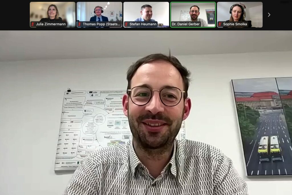Bildschirmfoto aus der Videokonferenz mit Daniel Gerber in der Hauptansicht. Darüber in Reihe als Miniaturansicht die Exptert:innen. Alle Teilnehmenden lächeln.